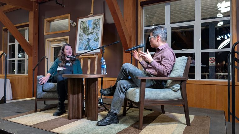 Two people sit on stage in armchairs, talking into microphones. A painting, water bottle, and large windows are in the background. The setting is warm, with wood beams and soft lighting.