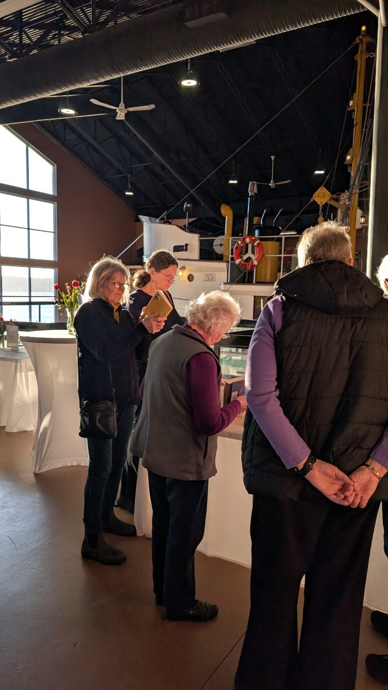 A group of older adults stands near a table at an indoor event with a boat displayed in the background. Sunlight streams through large windows, casting long shadows on the floor.
