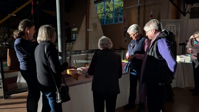 Five people stand around a table with stacked books, examining them in a brightly lit indoor space with nautical decor and large windows in the background.