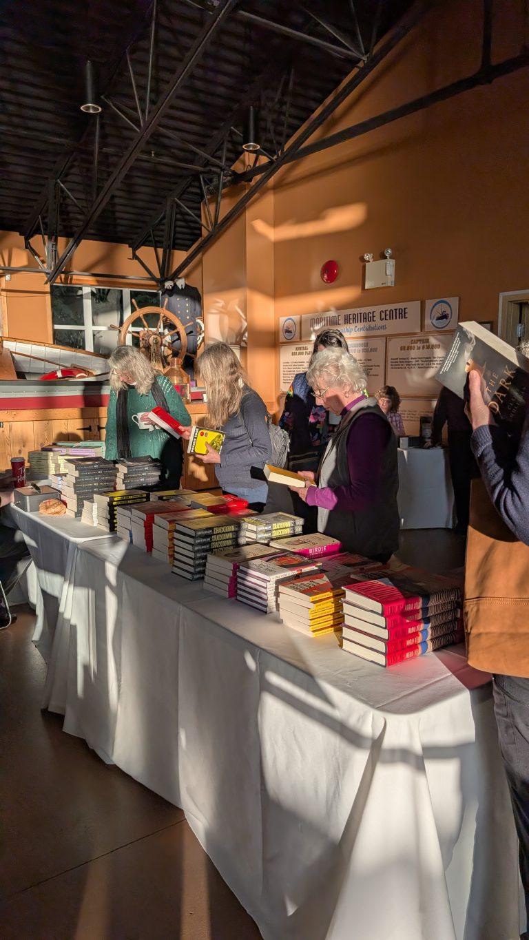 People browse stacks of books on a table inside a sunlit room with a vintage propeller plane and museum signs in the background.