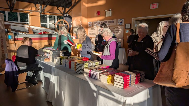 People browse and pick up books from stacks on a table at a well-lit indoor event. Most attendees have gray hair and are wearing jackets or sweaters. Sunlight streams through windows, casting long shadows across the scene.