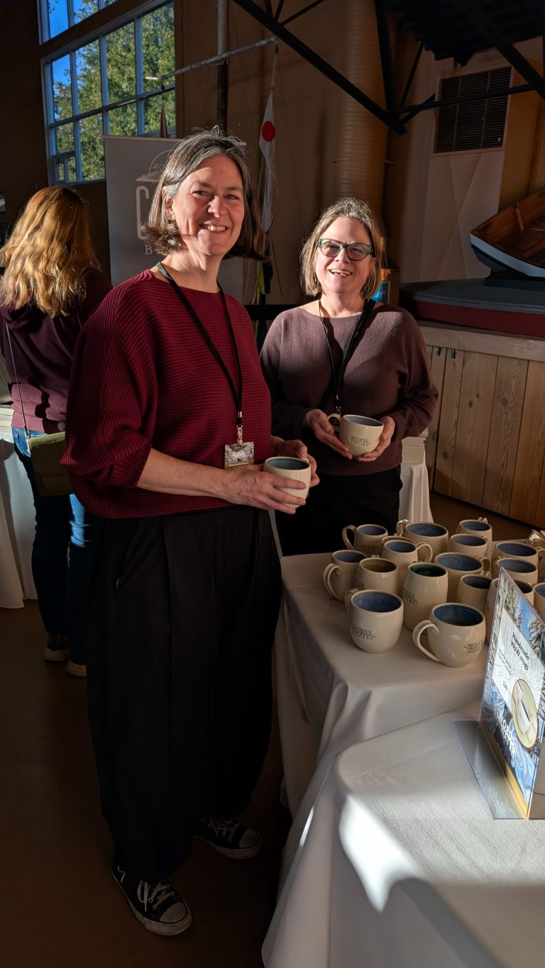 Two women stand by a table with mugs, smiling at the camera. Sunlight streams through windows behind them. Another person stands nearby facing away. The setting appears to be an indoor event or fair.