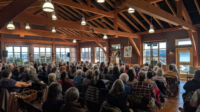 A large audience sits in a wooden hall with high rafters, listening to a speaker at a podium by the windows. Sunlight shines in, and there is a view of water outside.