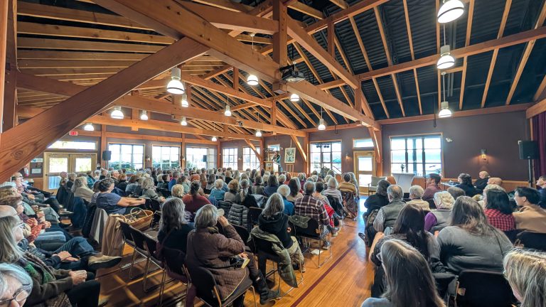 A large group of people sit in rows of chairs inside a wooden-beamed hall, facing a panel of speakers at the front of the room during a daytime event.