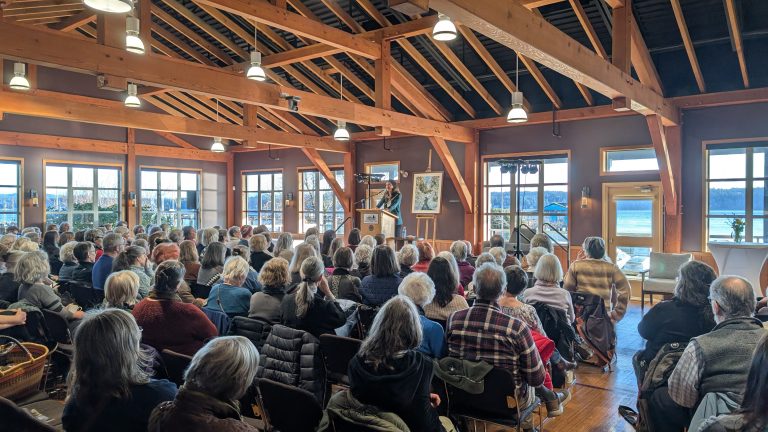 A large audience sits facing a speaker at a podium in a wooden hall with large windows, natural light, and scenic views outside.