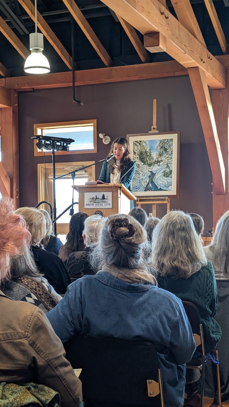 A woman speaks at a podium to an audience in a wooden room with exposed beams. A painting of a river scene is displayed behind her, and natural light comes in through the windows.