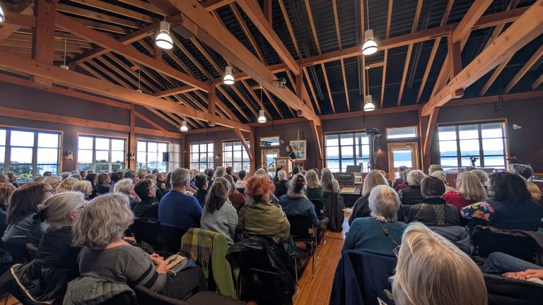 An audience of mostly older adults sits in a wooden-beamed room, facing a speaker at the front. The space has large windows and natural light.