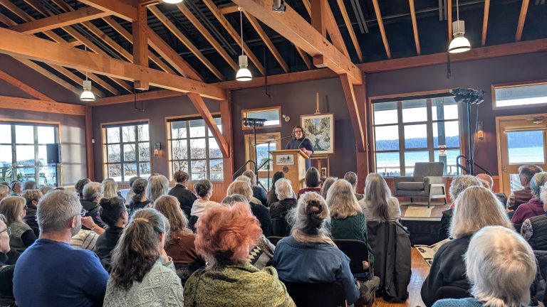 A speaker stands at a podium addressing a seated audience in a wooden-beamed room with large windows, artwork, and a view of water and trees outside.