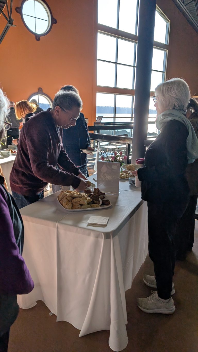 People gather around a table with a white tablecloth, where a man is serving bread. Sunlight streams through large windows in the background, creating a warm atmosphere.