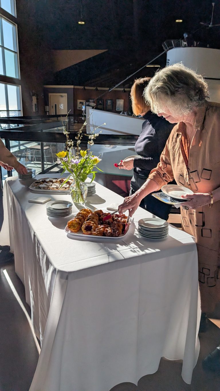 Three people serve themselves pastries from a table covered with a white cloth, plates, and a vase of flowers. Sunlight streams in through large windows, casting shadows on the floor.