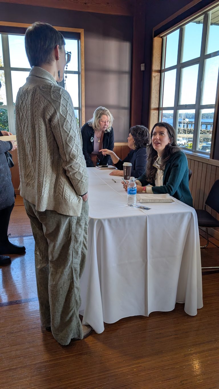 Two women sit at a table signing books and talking with visitors in a sunny room with large windows. One person stands in front of the table, wearing a cream sweater and green pants.