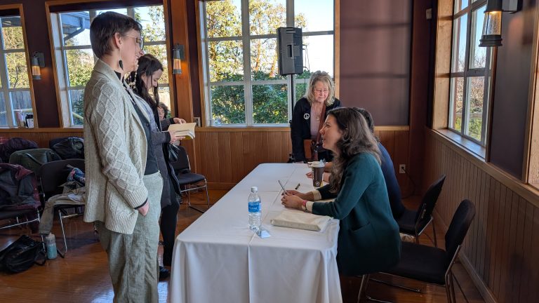 A woman sits at a table signing books while three people stand nearby, talking with her in a sunlit room with large windows and wooden walls.