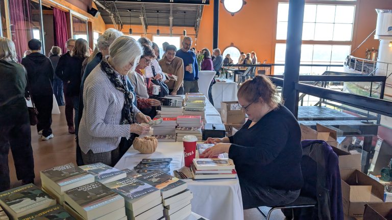 People line up at a book signing event, where an author sits at a table signing books. Stacks of books and a coffee cup are on the table. Sunlight streams in through large windows in the background.
