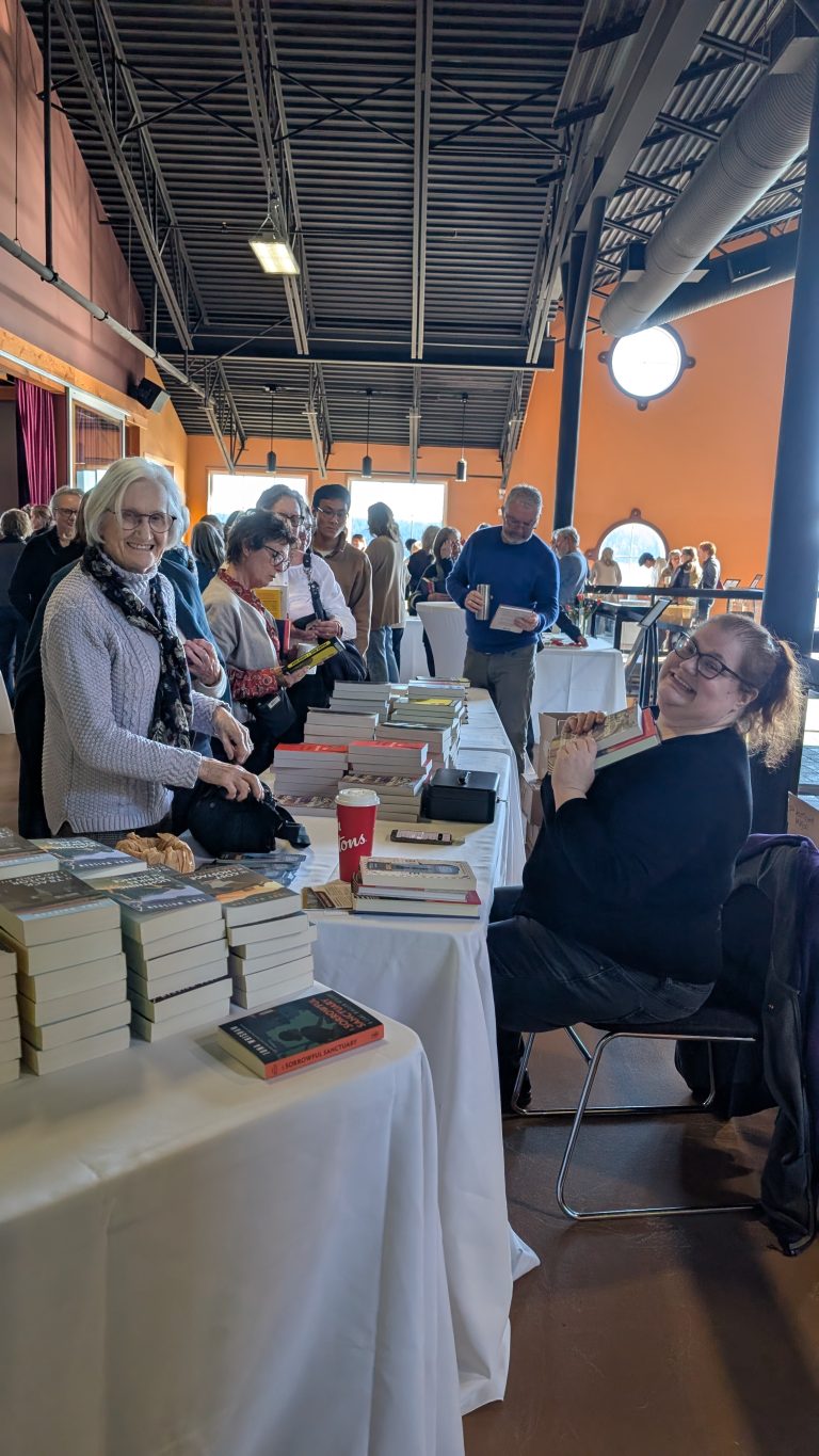 A group of people stand around a table stacked with books at an indoor event. One woman sits and smiles while holding books, as others interact with her and each other. The setting appears casual and friendly.