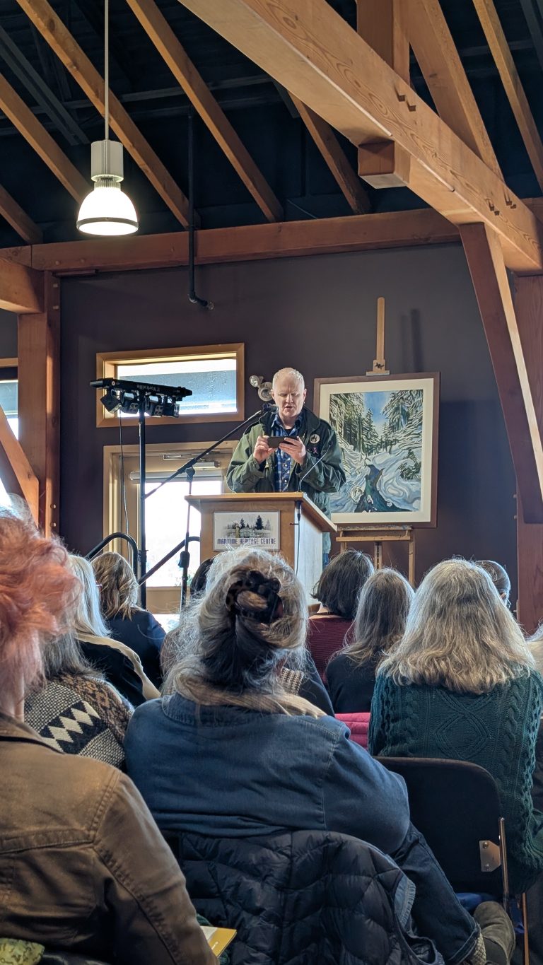 An older man speaks at a podium inside a wooden building, addressing a seated audience. Behind him is a painting of a river and forest, and lights hang from exposed beams above.