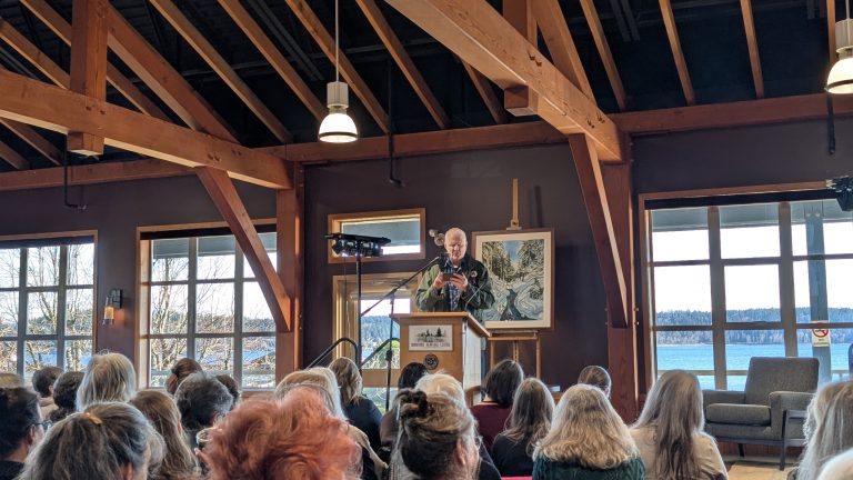 A speaker stands at a podium addressing an audience in a wooden hall with large windows overlooking water and trees. Framed art is displayed behind the podium.
