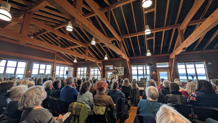 A large crowd sits in a rustic, wood-beamed hall facing a stage with a speaker at a podium. Large windows reveal a scenic outdoor view.