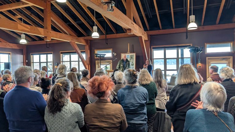 A crowd of people stands and faces a speaker at the front of a wooden-beamed room with large windows, applauding. The atmosphere is warm and filled with natural light.