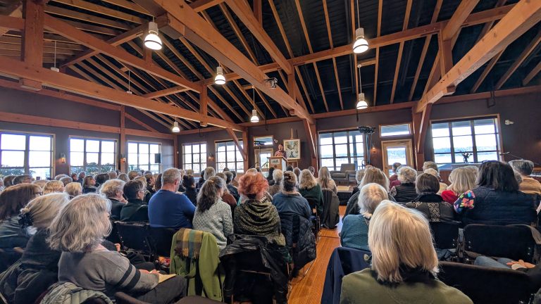 A large group of people sits in rows inside a wooden hall, facing a speaker at the front. Sunlight streams through the many windows, illuminating the audience and exposed wooden beams above.