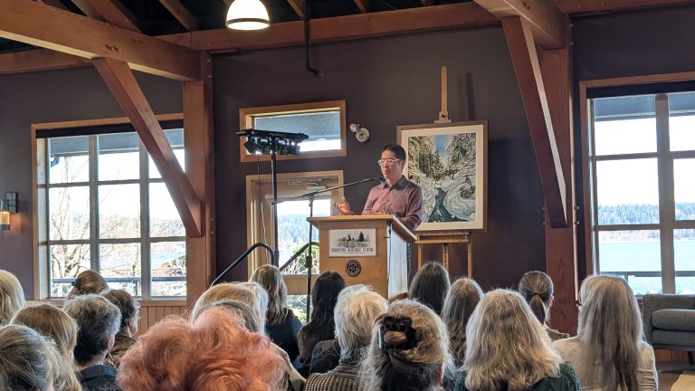A man speaks at a podium to an audience in a wood-beamed room with large windows and landscape art on display.