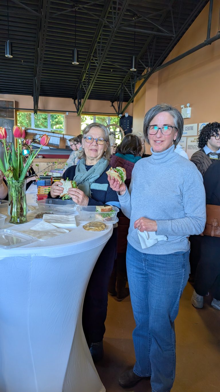 Two women stand at a tall table with flowers, smiling and holding sandwiches at a social event. Other people mingle in the background of the bright, indoor venue.