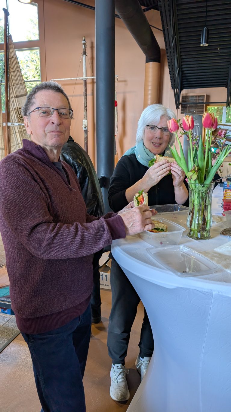 Two people stand at a tall table, eating sandwiches. The woman wears glasses and a scarf, the man wears glasses and a maroon sweater. A vase of pink tulips is on the table. Indoor setting with soft natural light.