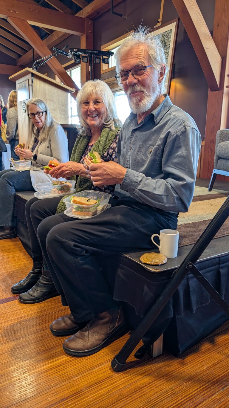 Three older adults sit indoors on benches and smile while eating lunch from plastic containers. A cookie and white mug sit on a chair nearby. Warm lighting and wooden beams are visible in the background.