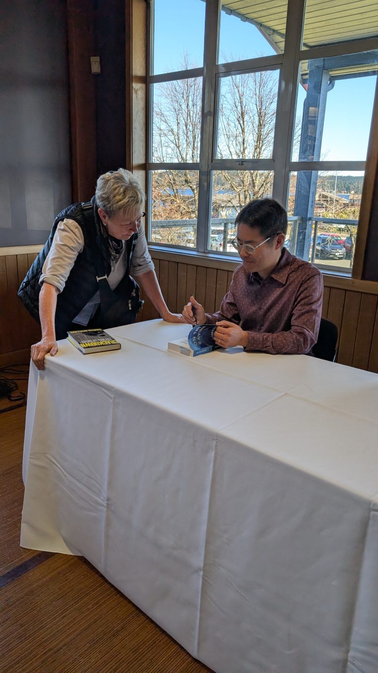 A man sits at a table signing a book while a woman stands beside him, leaning over and engaging in conversation. A book is on the table, and large windows show a sunny outdoor scene in the background.
