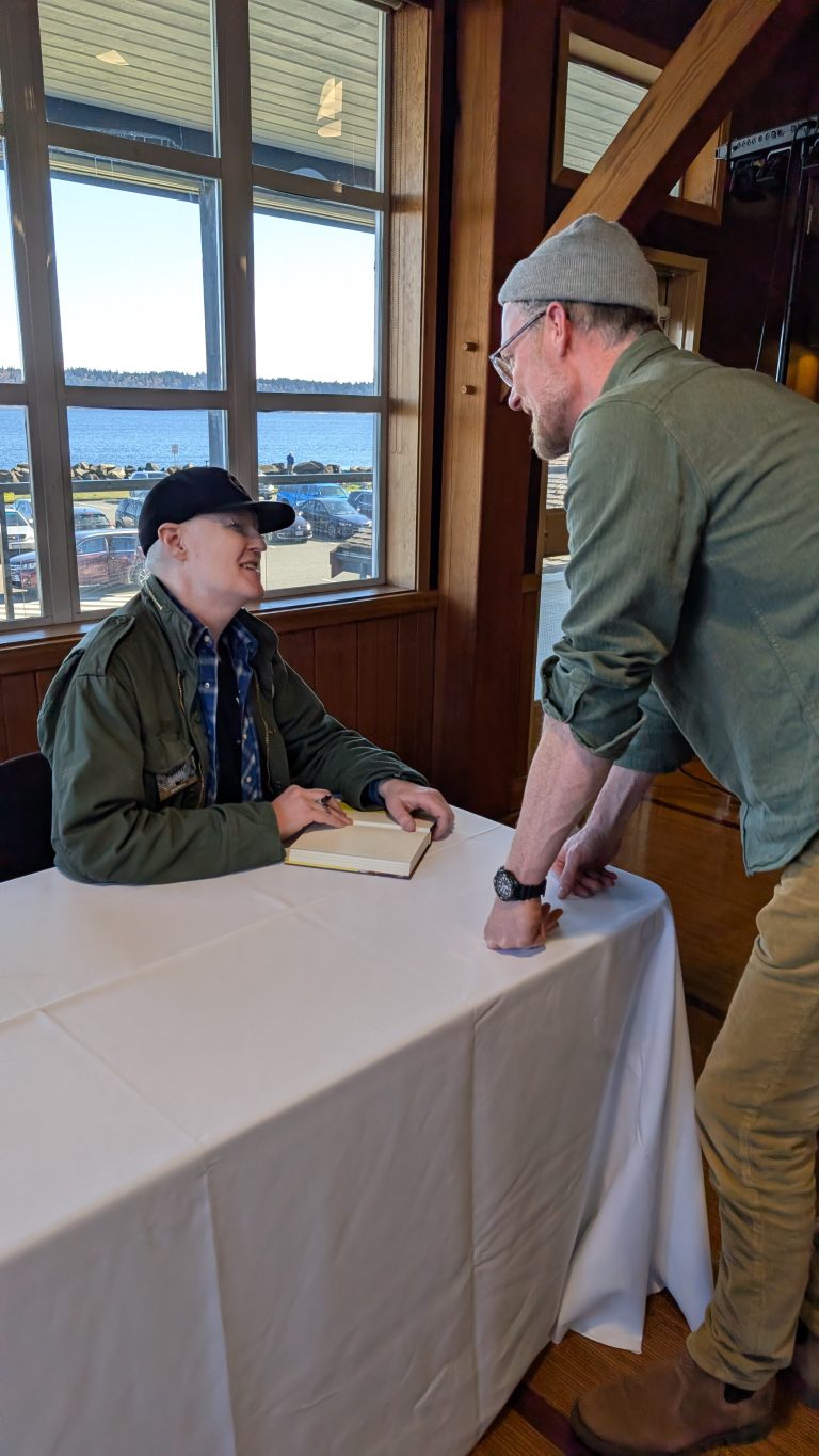Two people interact at a book signing event; one sits at a table with an open book and pen, smiling up at the other, who stands, leaning on the table, engaged in conversation. Large windows show a water view outside.