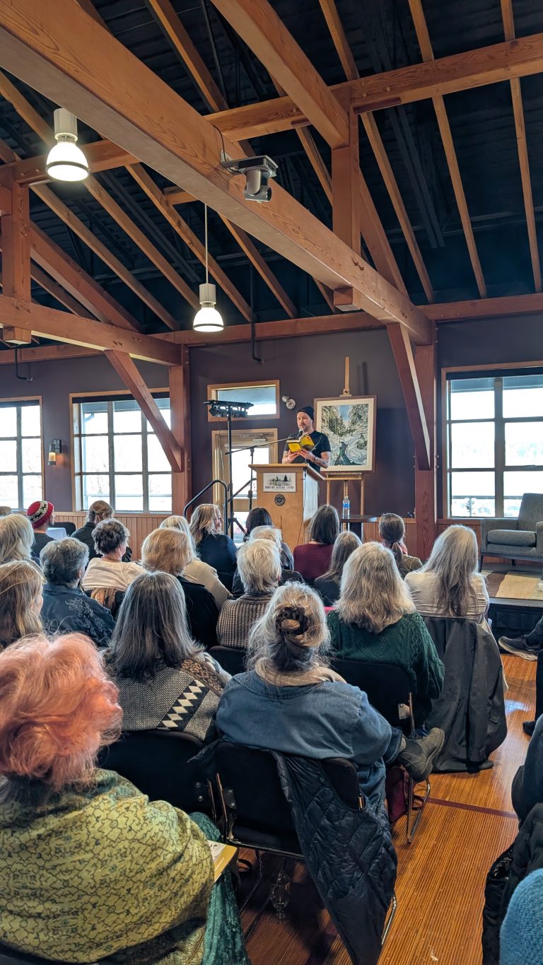 A speaker stands at a podium addressing an attentive audience in a room with exposed beams, wooden ceilings, and large windows letting in natural light.