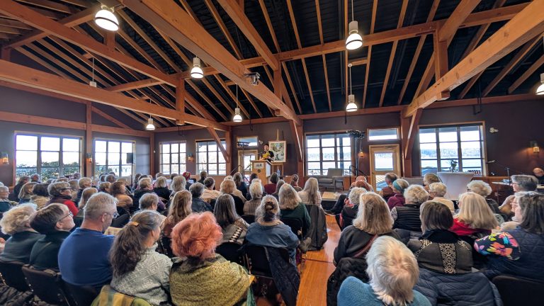 A large group of people sits facing a speaker at a podium inside a wooden-beamed hall with natural light coming from large windows.