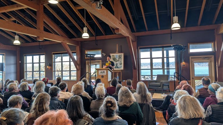A speaker addresses a seated audience in a wooden event hall with large windows showing a scenic view of water and trees outside.