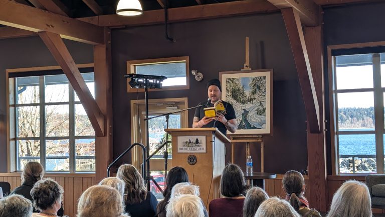 A person stands at a podium speaking to an audience in a wooden hall with large windows showing a lake view. The speaker holds a yellow vest and wears a black shirt. Artwork and a microphone are visible behind them.