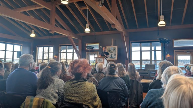 A speaker stands at a podium addressing a seated audience in a spacious, wooden-beamed room with large windows letting in natural light.