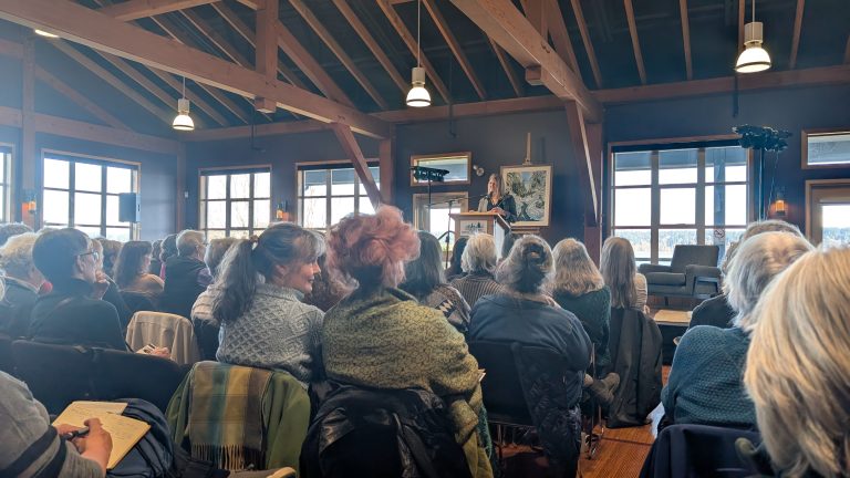 A woman speaks at a podium to a seated audience in a rustic, wooden-beamed room with large windows and natural light.