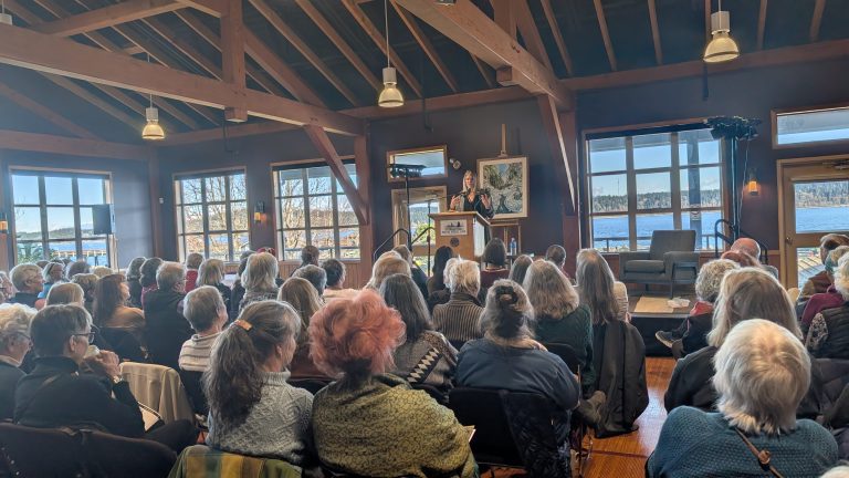A woman stands at a podium speaking to a seated audience in a rustic room with wooden beams, large windows, and scenic views of water and trees outside.