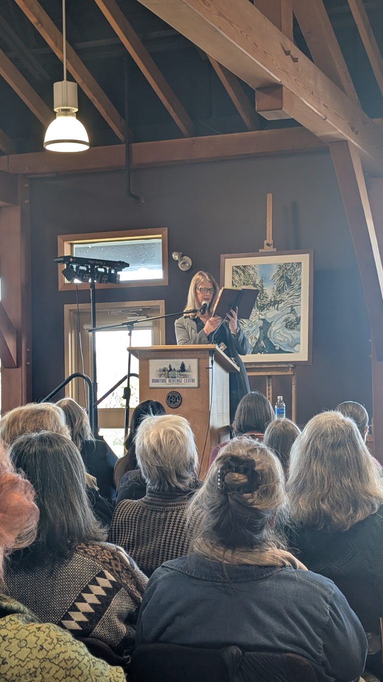 A woman speaks at a podium to an audience in a wooden room. Artwork hangs behind her, and sunlight enters through a window. The audience is seated and attentively listening.