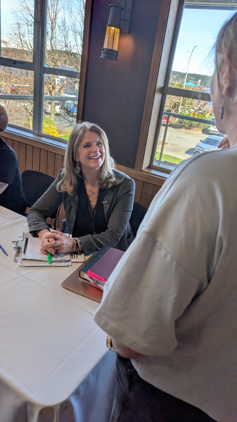 A woman sits at a table with papers and a highlighter, smiling and talking to another person standing nearby in a sunlit room with large windows.