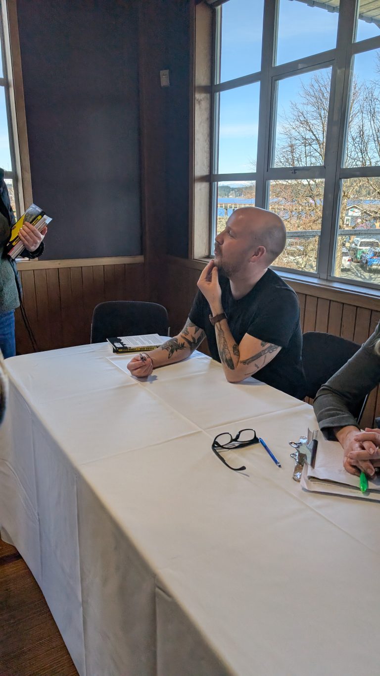 A person with tattoos and a beard sits at a table with a white tablecloth, eyeglasses, and a pen. They appear to be listening to someone standing nearby, with large windows and trees visible in the background.