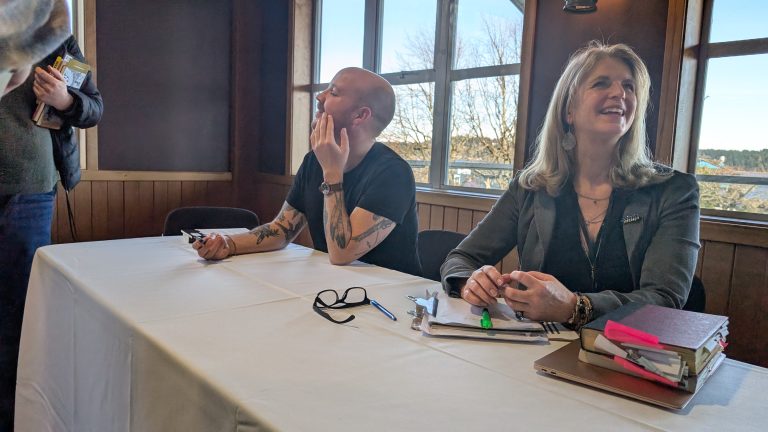 Two people sit at a table covered with a white cloth, one looking up while touching their face, the other smiling with a notepad, clipboard, and books in front of them. Sunlight streams through large windows behind them.