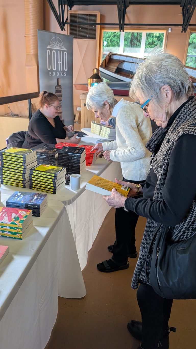 Three women browse books on a table at an indoor book fair. Stacks of colorful books are displayed, and a COHO Books banner stands in the background.