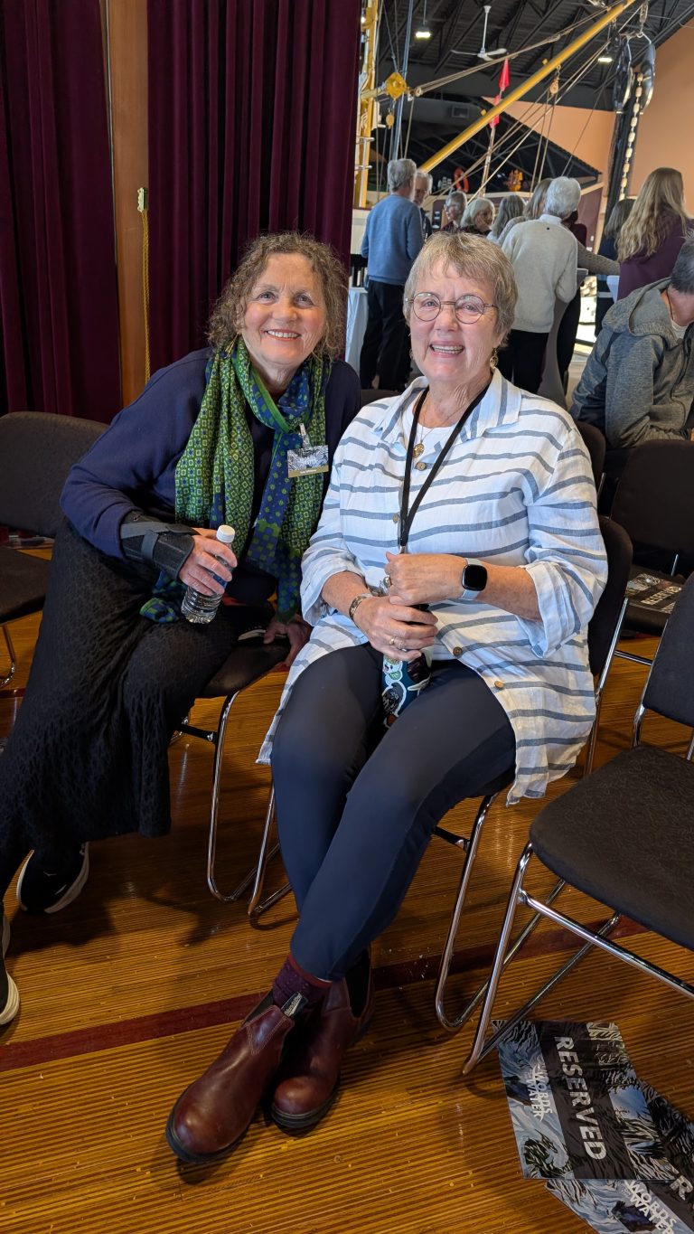 Two women sit and smile at an indoor event. One wears a striped shirt and glasses, the other has curly hair, a dark outfit, and a green scarf. People stand and talk in the background with chairs around them.