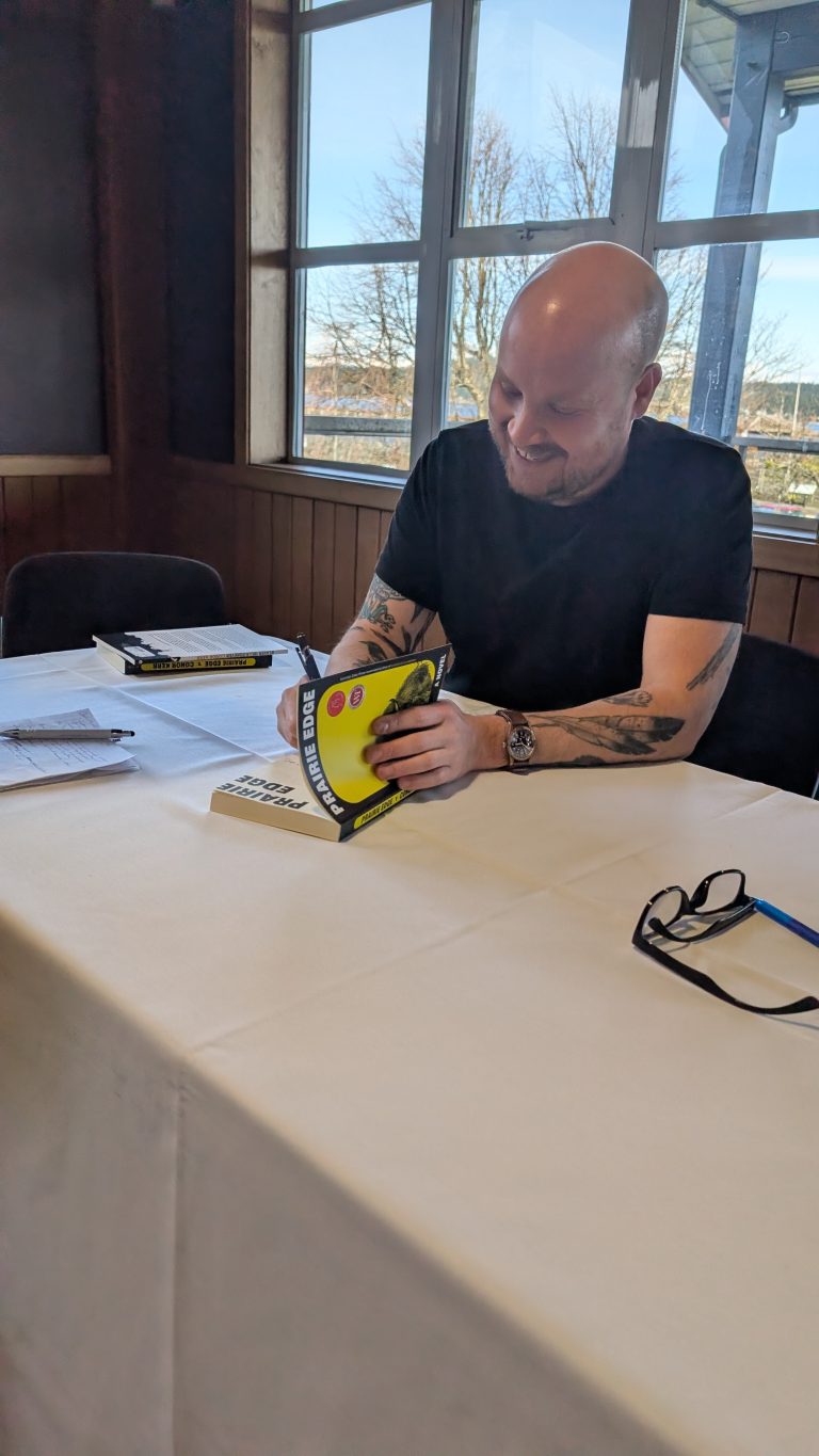 A smiling, bald man with tattoos sits at a table signing a yellow book. Papers, glasses, and another book are on the white tablecloth. Sunlight streams through large windows behind him.