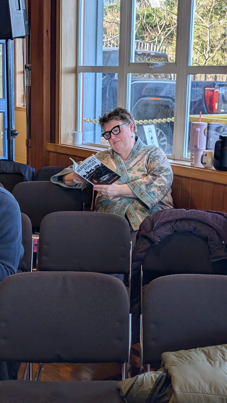 A person wearing glasses and a patterned jacket sits alone in a sunlit room, reading a magazine among empty chairs. A pink thermos and black kettle are on the windowsill behind them.