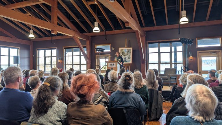 A group of people sit in a wooden-beamed room, attentively listening to a speaker at a podium in front of windows with scenic outdoor views.