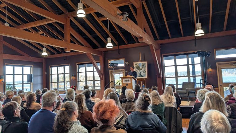 A speaker addresses a seated audience in a spacious, wooden-beamed room with large windows and natural light. People listen attentively, and a scenic view of water and trees is visible outside.