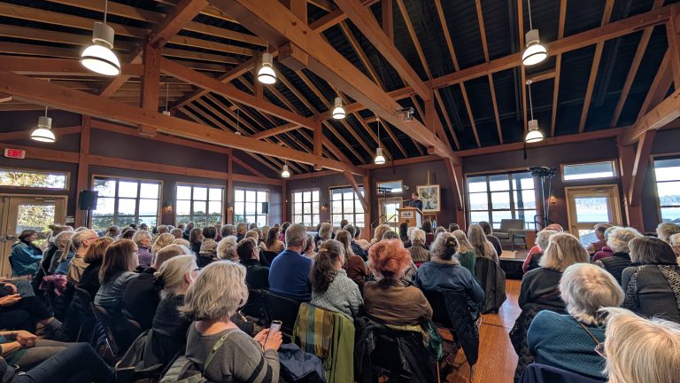 A large group of people sit in rows of chairs, attentively facing a speaker at the front of a wooden-beamed room with large windows letting in natural light.