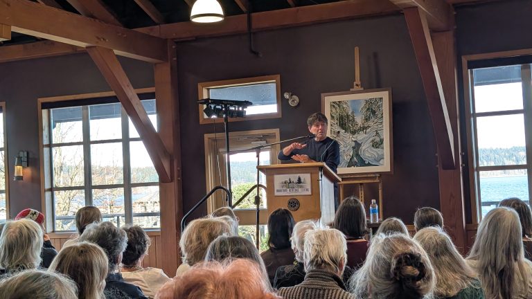 A person speaks at a podium in front of an audience in a room with large windows. Artwork and water views are visible in the background.