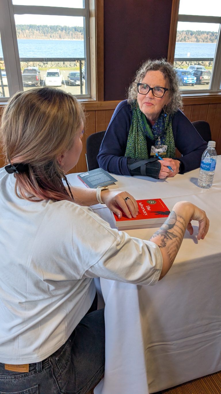 Two women sit across from each other at a table, talking. One woman holds a red book, while the other wears glasses, a green scarf, and a wrist brace. A water bottle sits on the table. Large windows are in the background.