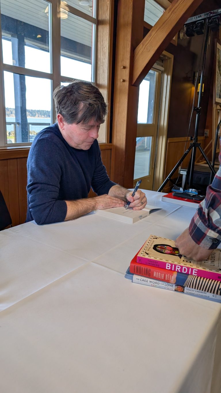A man with brown hair sits at a table signing a book. Several books are stacked nearby, and another person’s hands are visible holding a book. The setting appears to be a cozy indoor space with wooden walls and windows.